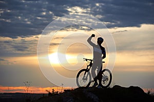 Sporty smiling man stands with bicycle on rock at top of hill against blurred background beautiful sunset