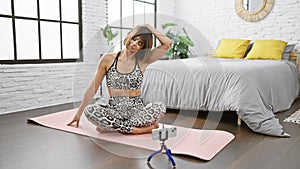 Sporty, smiling young hispanic woman beautifully records a yoga mat stretching tutorial, sitting in the bedroom of her warm indoor