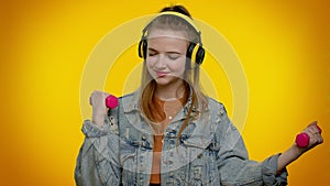 Sporty teen student girl listening music via headphones, working out, pumping up lifting dumbbells