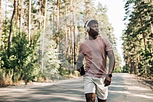 Sporty tired African American man runner, drinking water resting after training, on a city road in nature