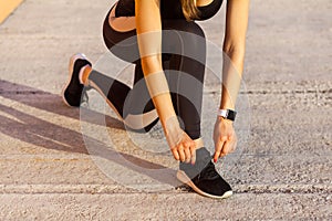 Sporty woman in black sporwear at morning on street standing on knee and preparing for training, tying shoelaces on sneakers