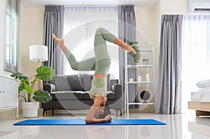 Sporty woman in green top and leggings standing in Salamba sirsasana exercise, Headstand pose on blue mat in bedroom at home