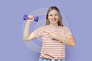 Sporty woman holding dumbbell in hand, female pointing at her biceps and smiling.