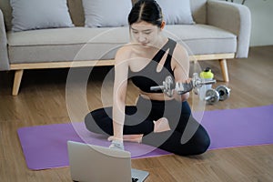 Sporty woman in sportswear is sitting on the floor with dumbbells and a protein shake or a bottle of water and is using a laptop