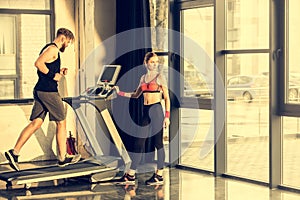 Sporty woman standing near bearded sportsman training on treadmill