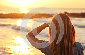 Sporty woman watches sunset over the sea, shading her eyes with one hand, view from behind only hair visible