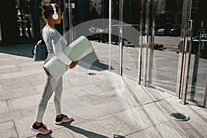 Sporty yoga woman in active wear and headphones walking with mat for workout