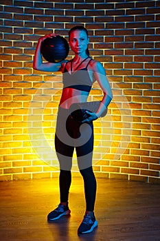 Sporty young brunette woman posing with medicine ball and foam fitness roller in gym in neon lights.