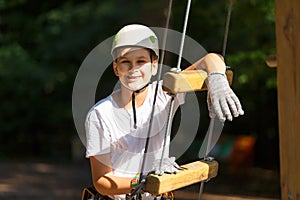 Sporty, young, cute boy in white t shirt spends his time in adventure rope park in helmet and safe equipment in the park