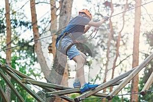 Sporty, young, cute boy in white t shirt spends his time in adventure rope park in helmet and safe equipment in the park