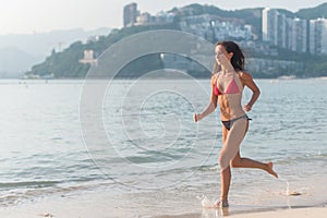 Sporty young woman in bikini running along the beach with bright sunlight and mountainous resort city in background