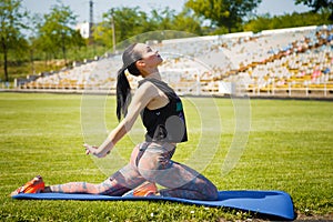 Sporty young woman stratching muscles on yoga mat on stadium green lawn. Sport, physical training, healthy way of life