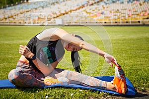 Sporty young woman stratching muscles on yoga mat on stadium green lawn. Sport physical training, healthy way of life, flexibility