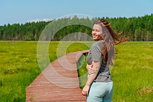 A sporty young woman walks along a picturesque wooden walking path through a swamp with tall grass in summer.