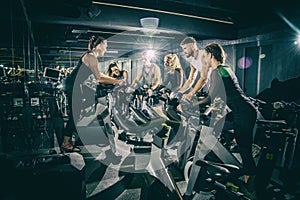 Sporty young women and men riding exercise bikes on cycling class with assistance of female instructor in the gym