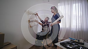 Spousal support, beautiful young wife and happy disabled husband in wheelchair choose wallpaper for room during
