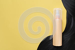 Spray bottle with thermal protection and lock of black hair on yellow background, flat lay. Space for text photo