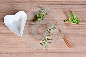 Sprigs of basil, mint and rosemary green aromatic flavor, empty white bowl in the shape of heart on wooden table