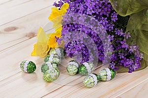 Spring bouquet of purple flowers and daffodils with chocolate eggs on a light wooden background