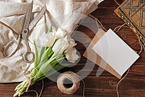 Spring bouquet of white tulip flowers, kraft envelope with blank card, scissors, twine on rustic wooden table. Wedding day composi