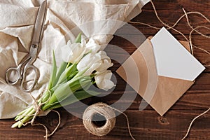 Spring bouquet of white tulip flowers, kraft envelope with blank card, scissors, twine on rustic wooden table. Wedding day composi