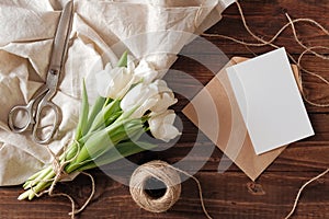Spring bouquet of white tulip flowers, kraft envelope with blank card, scissors, twine on rustic wooden table. Wedding day composi