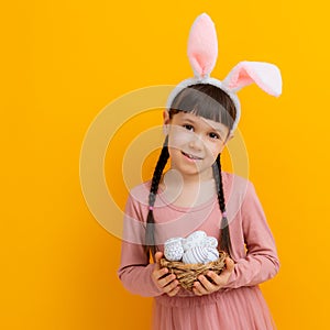 spring cheerful girl with rabbit ears on her head with basket colored eggs in her hands on a yellow background.