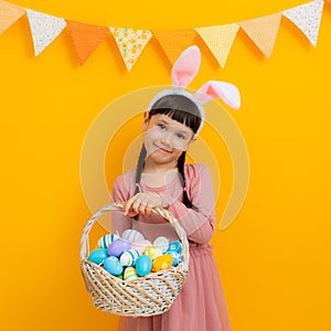 spring cheerful girl with rabbit ears on her head with basket colored eggs in her hands on a yellow background.