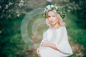Spring coming! Beautiful young cheerful pregnant woman in wreath flowers on head touching belly while walking in spring tree garde