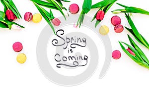 Spring is coming hand lettering surrounded by red tulips and sweets macarons on white background top view copy space