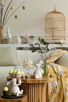 Spring composition of living room interior with round coffee table, beige sofa, easter bunny sculpture, colorful easter eggs,