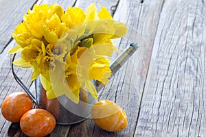 Spring daffodils in watering can with colorful eggs on wooden boards, easter decoration concept