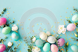 Spring Easter background with pastel easter eggs, flowers and eucalyptus decorated golden confetti on blue table. Top view photo