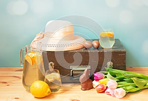 Spring and easter travel concept. Vintage wooden suitcase, hat, chocolate eggs, chocolate bunny, lemonade and tulip flowers