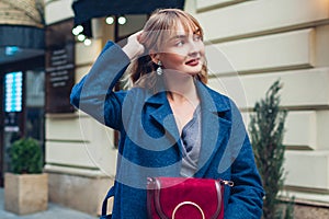 Spring fashion female accessories. Stylish young woman wearing trendy blue coat jewellery walking with purse on street.