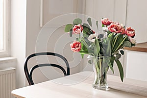 Spring flowers in glass vase on wooden table. Blurred kitchen background with old chair. Bouquet of red tulips, white