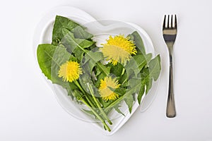 Spring fresh green salad with dandelions and edible flowers on a heart-shaped plate on white background. The concept of diet and