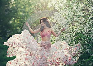 Spring girl with wavy hair dancing on a background of flowering trees. She wears a pink dress with flowers that flutters