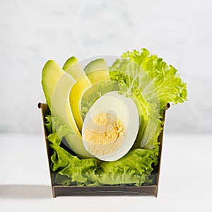 Spring green salad with lettuce, avocado, egg in paper bowl cut in half on light white wood table, marble tile wall, closeup.