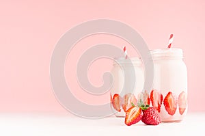 Spring healthy milk beverages with cut ripe strawberry, red striped straws on gentle pastel pink background, white wood table.