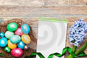 Spring holiday greeting card on Easter with empty notebook for recipe, nest with colorful eggs and flowers on rustic wooden table.