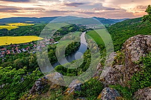 Jarní krajina s rozkvetlými poli, zelenými loukami a klikatící se řekou v údolí pod skalami.