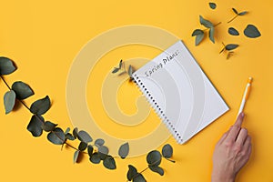 Spring plans text. Eucalyptus leaves and a notebook, a woman's hand writes with a pen. Yellow background.