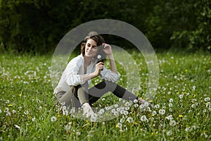 Spring portrait of a girl sitting in a field on the grass among dandelion flowers. Cheerful girl enjoys Sunny spring weather.
