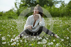 Spring portrait of a girl sitting in a field on the grass among dandelion flowers. Cheerful girl enjoys Sunny spring weather.