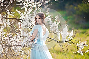 Spring touch. Happy beautiful young woman in blue dress enjoy fresh flowers and sun light in blossom park at sunset.