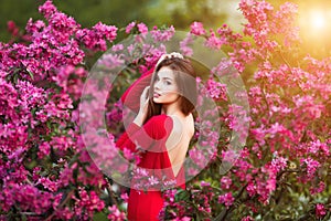 Spring touch. Happy beautiful young woman in red dress enjoy fresh pink flowers and sun light in blossom park at sunset.