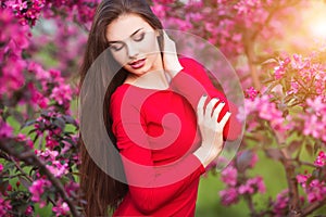 Spring touch. Happy beautiful young woman in red dress enjoy fresh pink flowers and sun light in blossom park at sunset.