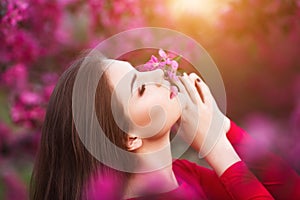 Spring touch. Happy beautiful young woman in red dress enjoy fresh pink flowers and sun light in blossom park at sunset.