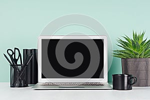 Spring workplace with blank notebook screen, black stationery, books, aloe plant in ribbed pot, coffee cup, in elegant green mint.